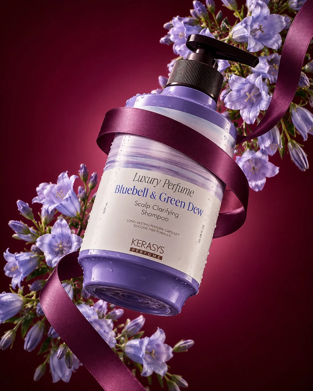 Shampoo bottle tilted diagonally on a burgundy background, wrapped in a plum ribbon with lilac bellflowers