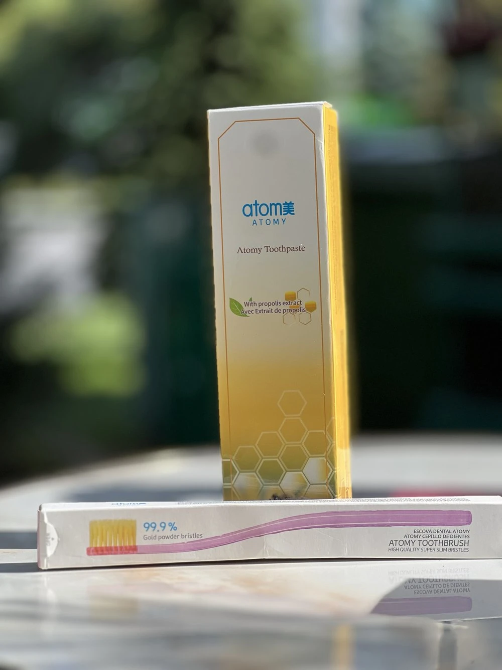 Original phone photo of a toothpaste box and a toothbrush box outdoors with a blurred green background